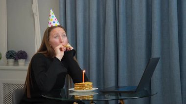 Renkli parti şapkalı genç bir kadın doğum gününü neredeyse kutluyor. Pastanın tadını çıkarırken bayram kornası çalıyor ve dizüstü bilgisayarından ev mutfağından video arıyor..