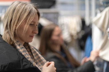 Şık siyah ceket ve desenli eşarp giymiş sarışın bir kadın, bir moda mağazasında şık kıyafetler seçiyor, satın almadan önce seçeneklerini dikkatlice değerlendiriyor.