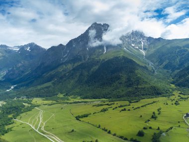 Nefes kesen hava manzarası girdaplı bulutlarla çevrili görkemli bir dağ zirvesini yakalıyor, kıvrımlı toprak yolları olan yemyeşil bir vadiye ve Svaneti, Georgia 'daki dağınık ağaçlara bakıyor.