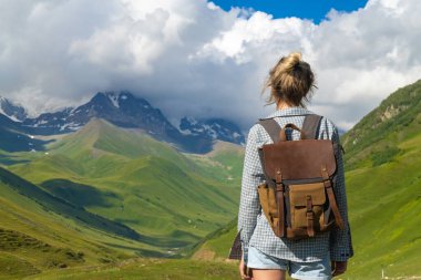 Sırt çantalı kadın yürüyüşçü, yeşil vadi ve karlı tepelerin nefes kesici manzarasının keyfini çıkarıyor. Dağ macerası sırasında doğanın güzelliğini kucaklıyor.