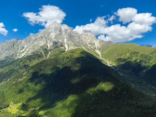 Görkemli bir dağ zirvesinin büyüleyici panoramik görüntüsü açık mavi gökyüzünün altında parlak yeşil bir vadi üzerine uzun gölgeler bırakıyor Svaneti, Georgia 'da tüylü bulutlar var.