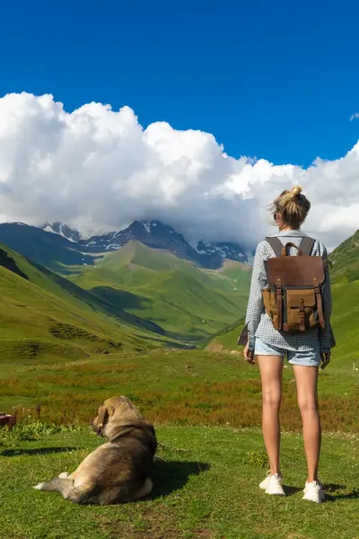 Sırt çantalı kadın yürüyüşçü köpeğiyle çimenli yamaçta duruyor, Ushguli, Gürcistan 'da büyüleyici manzaranın tadını çıkarıyor, görkemli dağlarla ve beyaz bulutlu mavi gökyüzüyle çevrili. Dikey fotoğraf