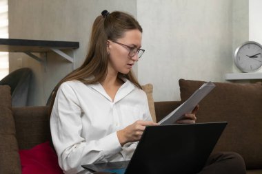 Gözlüklü genç iş kadını rahat bir koltukta oturuyor, dizüstü bilgisayarla çalışıyor, önemli belgeleri dikkatlice okuyor ve analiz ediyor.