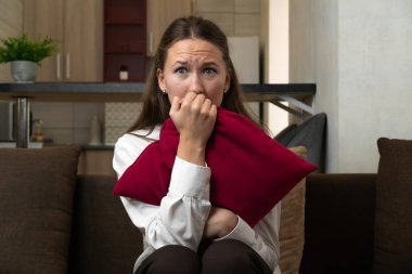 Evdeki kanepede oturan genç bir kadın yoğun korku ve endişe yaşıyor, kırmızı bir yastığı sıkıca tutuyor ve tırnaklarını yiyor, bir anlık kırılganlık ve stres gösteriyor.