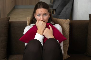 Endişeli genç bir kadın evindeki koltukta otururken parmaklarını ısırıyor ve kırmızı bir yastığa sarılıyor. Korku, endişe ve stresi ifade ediyor.