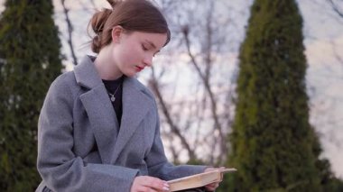 Genç kız öğrenci parkta bankta dinleniyor, kitap okuyor ve kahve içiyor, yeşil bölgede güneşli ve huzurlu bir bahar gününün tadını çıkarıyor.