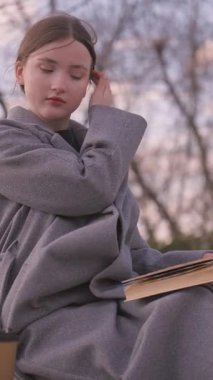 Gri montlu genç kız öğrenci parkta bir bankta oturmuş kitap okuyor ara sıra sayfaları çeviriyor, ara sıra esniyor, dışarıda doğa ile çevrili sakin bir zaman geçiriyor.