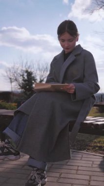 Gri montlu genç kız öğrenci parkta bir bankta oturmuş kitap okuyor ara sıra sayfaları çeviriyor, ara sıra esniyor, dışarıda doğa ile çevrili sakin bir zaman geçiriyor.