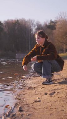 Genç bir kadın göl kıyısında çömelmiş, ekmeğini nazikçe yiyor ve güneşli bahar günü dikey videosu boyunca huzurlu ve sakin bir tavırla yüzen ördekleri besliyor.
