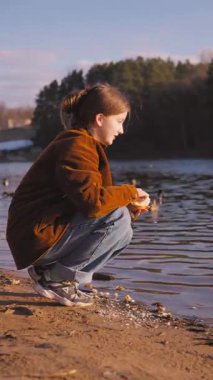 Genç bir kadın göl kıyısında çömelmiş, ekmeğini nazikçe yiyor ve güneşli bahar günü dikey videosu boyunca huzurlu ve sakin bir tavırla yüzen ördekleri besliyor.