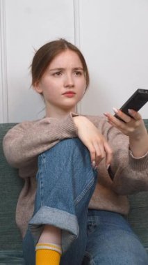 Genç kız kanepede dinleniyor, uzaktan kumandayı tutuyor ve yüz ifadesini değiştirerek heyecan verici dikey video arayışı içinde.