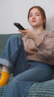 Genç kız kanepede dinleniyor, uzaktan kumandayı tutuyor ve yüz ifadesini değiştirerek heyecan verici dikey video arayışı içinde.