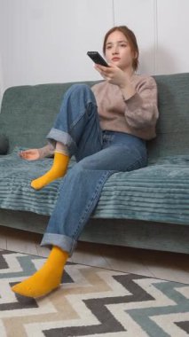 Genç kız kanepede dinleniyor, uzaktan kumandayı tutuyor ve yüz ifadesini değiştirerek heyecan verici dikey video arayışı içinde.