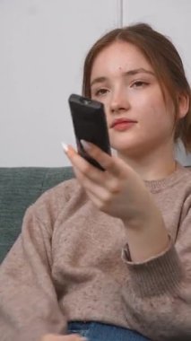 Rahat genç kız kablosuz uzaktan kumandayı göstererek rahat koltukta dinleniyor, TV kanalları izliyor ve evde dikey videonun keyfini çıkarıyor.