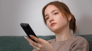 Rahat genç kız kablosuz uzaktan kumandayı göstererek rahat koltukta dinleniyor, TV kanalları izliyor ve evde eğleniyor.