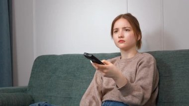 Rahat genç kız kablosuz uzaktan kumandayı göstererek rahat koltukta dinleniyor, TV kanalları izliyor ve evde eğleniyor.