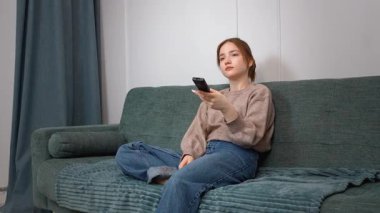 Rahat genç kız kablosuz uzaktan kumandayı göstererek rahat koltukta dinleniyor, TV kanalları izliyor ve evde eğleniyor.