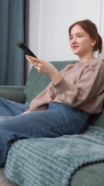 Rahat genç kız kablosuz uzaktan kumandayı göstererek rahat koltukta dinleniyor, TV kanalları izliyor ve evde dikey videonun keyfini çıkarıyor.