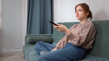 Rahat genç kız kablosuz uzaktan kumandayı göstererek rahat koltukta dinleniyor, TV kanalları izliyor ve evde eğleniyor.