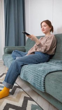 Oturma odasındaki kanepede oturan güzel genç kız uzaktan kumandayı kullanıyor, kanal değiştiriyor, ev ortamında televizyon programlarının keyfini çıkarıyor.
