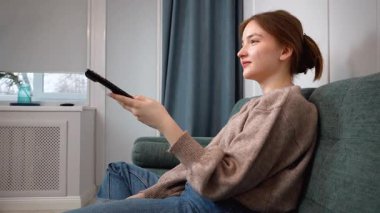Oturma odasındaki kanepede oturan güzel genç kız uzaktan kumandayı kullanıyor, kanal değiştiriyor, rahat bir ev ortamında televizyon programlarının keyfini çıkarıyor.