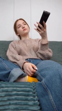 Oturma odasındaki kanepede oturan güzel genç kız uzaktan kumandayı kullanıyor, kanal değiştiriyor, ev ortamında televizyon programlarının keyfini çıkarıyor.