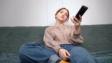 Oturma odasındaki kanepede oturan güzel genç kız uzaktan kumandayı kullanıyor, kanal değiştiriyor, rahat bir ev ortamında televizyon programlarının keyfini çıkarıyor.