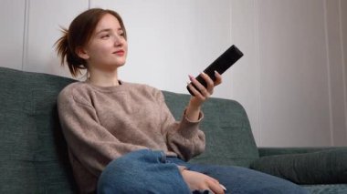 Oturma odasındaki kanepede oturan güzel genç kız uzaktan kumandayı kullanıyor, kanal değiştiriyor, rahat bir ev ortamında televizyon programlarının keyfini çıkarıyor.