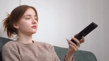 Rahat genç bir kız rahat bir koltukta oturuyor TV kanallarını değiştiriyor evinde kablosuz uzaktan kumandayla eğleniyor.