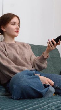 Rahat bir genç kız rahat bir koltukta oturuyor TV kanallarını değiştiriyor evinde kablosuz uzaktan kumandalı dikey video ile eğleniyor.