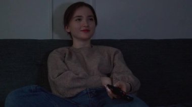 Rahat genç kız boş zamanlarını kanepede oturup televizyon izleyerek geçirmeyi ve loş ışıklı oturma odasındaki kanalı değiştirmek için uzaktan kumandayı kullanmayı seviyor.