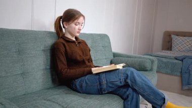 Öğrenci kız rahat bir şekilde kitap okuyarak, sıcak bir oturma odasındaki yumuşak bir kanepede oturarak, kişisel rahatlama ve edebi araştırma anının tadını çıkararak dinlendi.