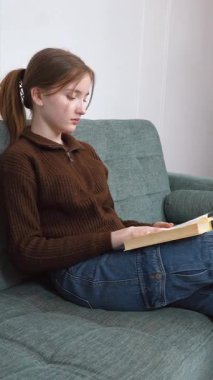 Öğrenci kız rahat bir şekilde kitap okuyarak, sıcak bir oturma odasındaki yumuşak bir kanepede oturarak, kişisel rahatlama ve edebi araştırma dikey videosunun keyfini çıkararak dinlendi.