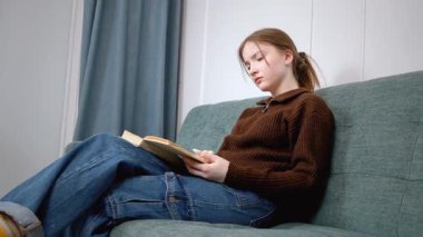 Öğrenci kız rahat bir şekilde kitap okuyarak, sıcak bir oturma odasındaki yumuşak bir kanepede oturarak, kişisel rahatlama ve edebi araştırma anının tadını çıkararak dinlendi.