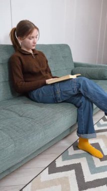 Öğrenci kız rahat bir şekilde kitap okuyarak, sıcak bir oturma odasındaki yumuşak bir kanepede oturarak, kişisel rahatlama ve edebi araştırma dikey videosunun keyfini çıkararak dinlendi.