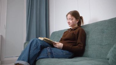 Öğrenci kız rahat bir şekilde kitap okuyarak, sıcak bir oturma odasındaki yumuşak bir kanepede oturarak, kişisel rahatlama ve edebi araştırma anının tadını çıkararak dinlendi.