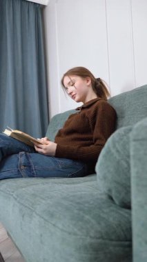 Genç kız öğrenci kitap okuyor. Edebiyata gömülmüş ve Minimalist Ev İçi Dikey Video Konsantrasyonu ile rahatça oturuyor.