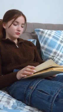 Edebiyata dalmış, rahat bir kanepede kareli yastıkların arasına rahatça oturmuş sakin bir ev keyfi ve kişisel sessiz zaman videosunu kişileştiren özgür genç bir kadın.