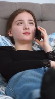 Genç bir kadın rahat kıyafetlerle rahat bir şekilde yatakta yumuşak yastıklarla uzanıyor, evde dikey videoda dinlenirken akıllı bir ifadeyle telefonda konuşuyor.