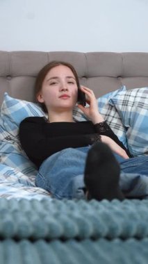 Genç bir kadın rahat kıyafetlerle rahat bir şekilde yatakta yumuşak yastıklarla uzanıyor, evde dikey videoda dinlenirken akıllı bir ifadeyle telefonda konuşuyor.