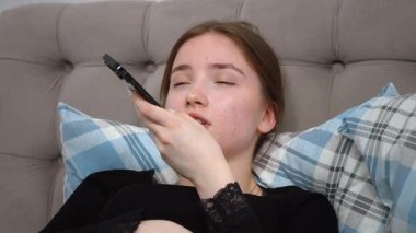 Rahat genç bir kadın, rahat bir şekilde yatakta uzanıyor, akıllı telefondan sesli mesaj kaydediyor, sakin bir dijital bağlantı ve rahatlama anının tadını çıkarıyor.