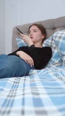 Rahatlamış genç kadın, rahat bir şekilde yatakta uzanıyor, akıllı telefondan sesli mesaj kaydediyor, sakin bir dijital bağlantı anının ve rahatlama dikey videosunun tadını çıkarıyor.