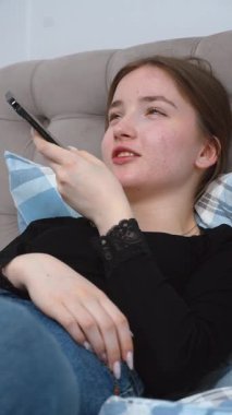 Rahatlamış genç kadın, rahat bir şekilde yatakta uzanıyor, akıllı telefondan sesli mesaj kaydediyor, sakin bir dijital bağlantı anının ve rahatlama dikey videosunun tadını çıkarıyor.