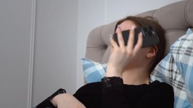 Rahat genç bir kadın, rahat bir şekilde yatakta uzanıyor, akıllı telefondan sesli mesaj kaydediyor, sakin bir dijital bağlantı ve rahatlama anının tadını çıkarıyor.