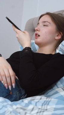 Rahatlamış genç kadın, rahat bir şekilde yatakta uzanıyor, akıllı telefondan sesli mesaj kaydediyor, sakin bir dijital bağlantı anının ve rahatlama dikey videosunun tadını çıkarıyor.