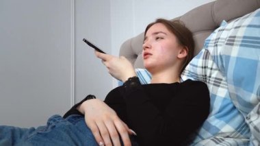 Genç bir kadın bir yastıkla yatakta uzanıyor, akıllı telefonunu kullanıyor, kulağına tutmakla, dinlemeyi önermek ve uzak tutmak arasında değişiyor, konuşmayı veya hoparlörü kullanmayı öneriyor.