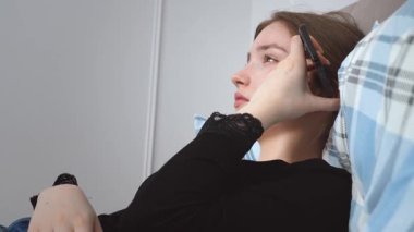 Genç bir kadın bir yastıkla yatakta uzanıyor, akıllı telefonunu kullanıyor, kulağına tutmakla, dinlemeyi önermek ve uzak tutmak arasında değişiyor, konuşmayı veya hoparlörü kullanmayı öneriyor.