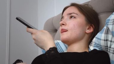 Genç bir kadın bir yastıkla yatakta uzanıyor, akıllı telefonunu kullanıyor, kulağına tutmakla, dinlemeyi önermek ve uzak tutmak arasında değişiyor, konuşmayı veya hoparlörü kullanmayı öneriyor.