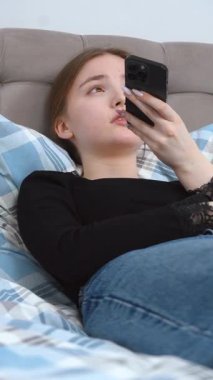 Rahat giyinen, yatakta dinlenen, sosyal medyada gezinirken akıllı telefon kullanan, mesajlaşan ve rahat ev ortamında dijital içerikle ilgilenen bir kadın.