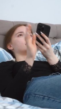 Rahat giyinen, yatakta dinlenen, sosyal medyada gezinirken akıllı telefon kullanan, mesajlaşan ve rahat ev ortamında dijital içerikle ilgilenen bir kadın.